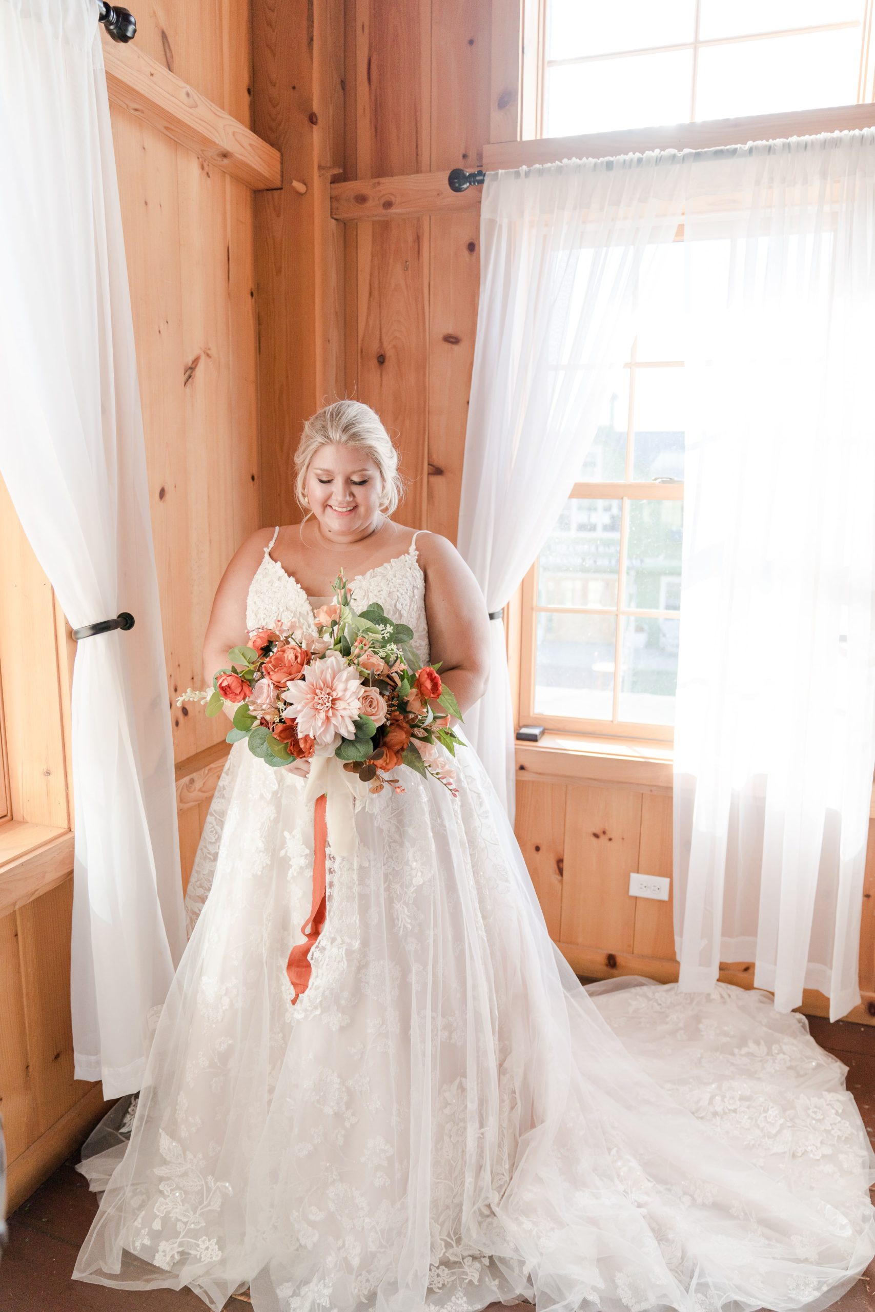 Picture of a bride standing with bouquet of flowers in her wedding dress in front of a window.